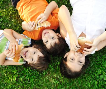 Three children laying on green grass on ground and eating sandwiches a